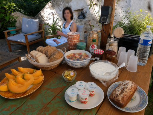 Le petit-déjeuner Table de repas extérieure avec melons, yaourt, confiture, pains et boissons, femme assise en arrière-plan.